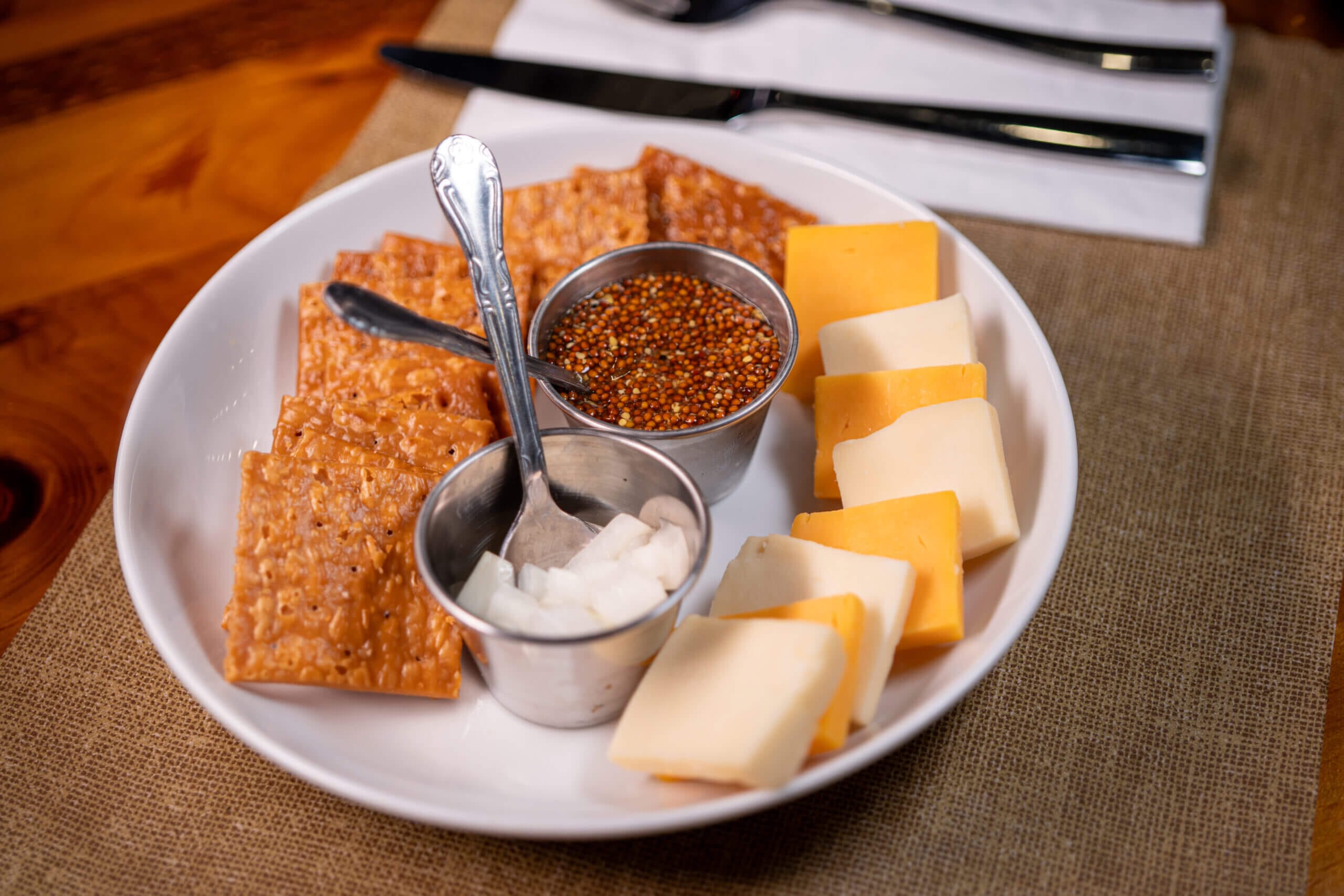 Cheese platter with assorted cheeses, crackers, pickled onions, and grainy mustard on a rustic table setting.