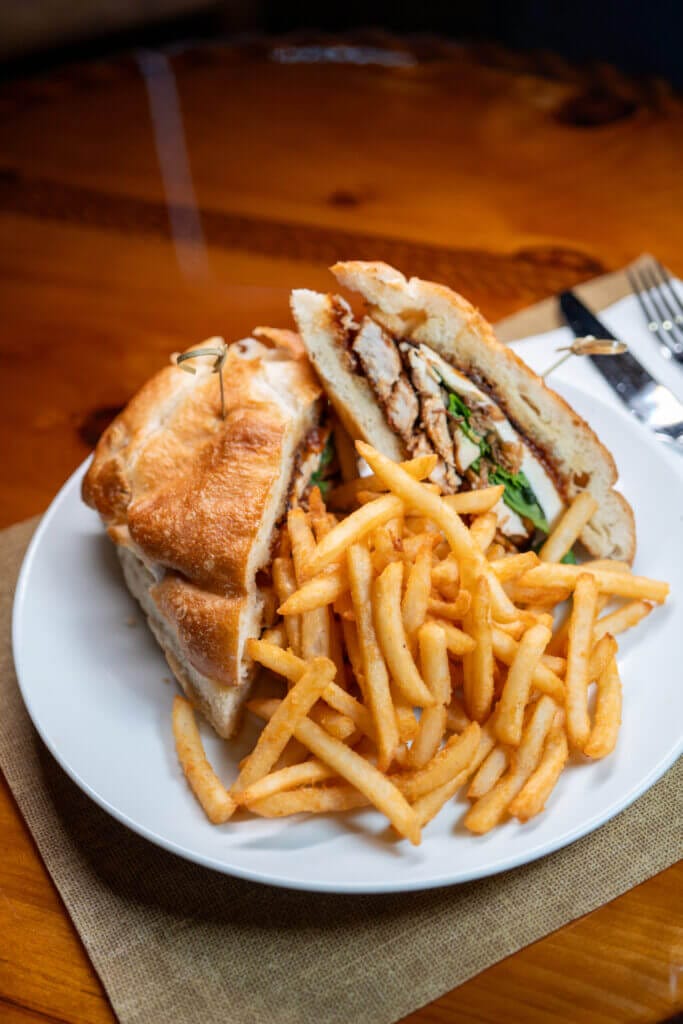 Chicken sandwich with fresh lettuce and tomato served with crispy golden french fries on a white plate on a wooden table.