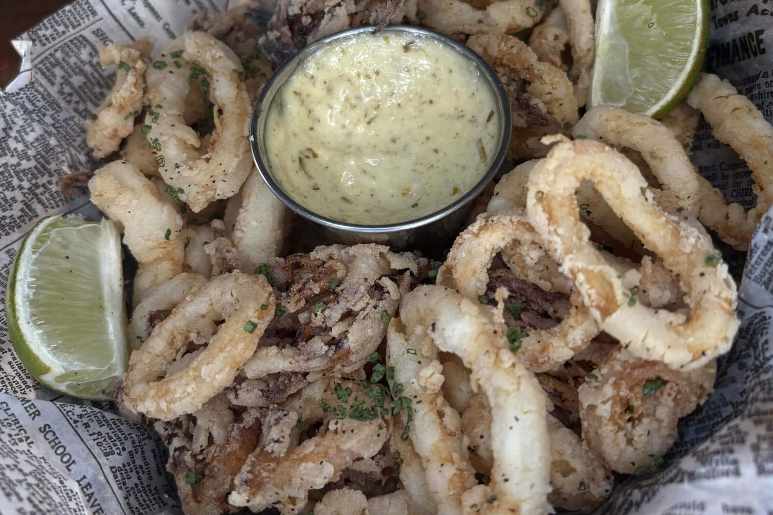 Crispy fried calamari with lime wedges and creamy dipping sauce on newspaper-lined plate.